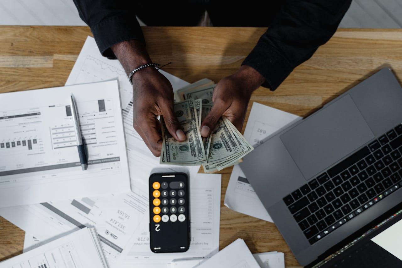 Services Person counting US dollars, using a calculator and laptop, with financial documents on a wooden desk.